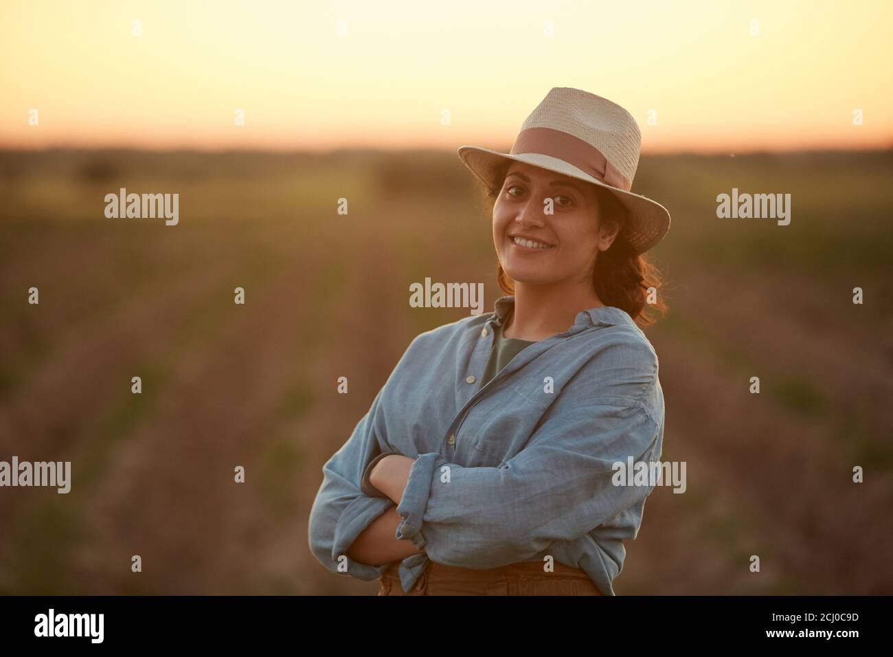 Waist-up-Porträt einer jungen Farmerin, die selbstbewusst mit gekreuzten Armen posiert, während sie bei Sonnenuntergang im Feld steht und die Kamera anlächelt, Raum kopieren Stockfoto