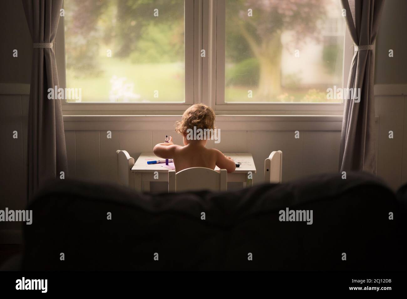 Kleinkind sitzt an einem weißen Schreibtisch vor den Fenstern Färben mit Markierungen Stockfoto