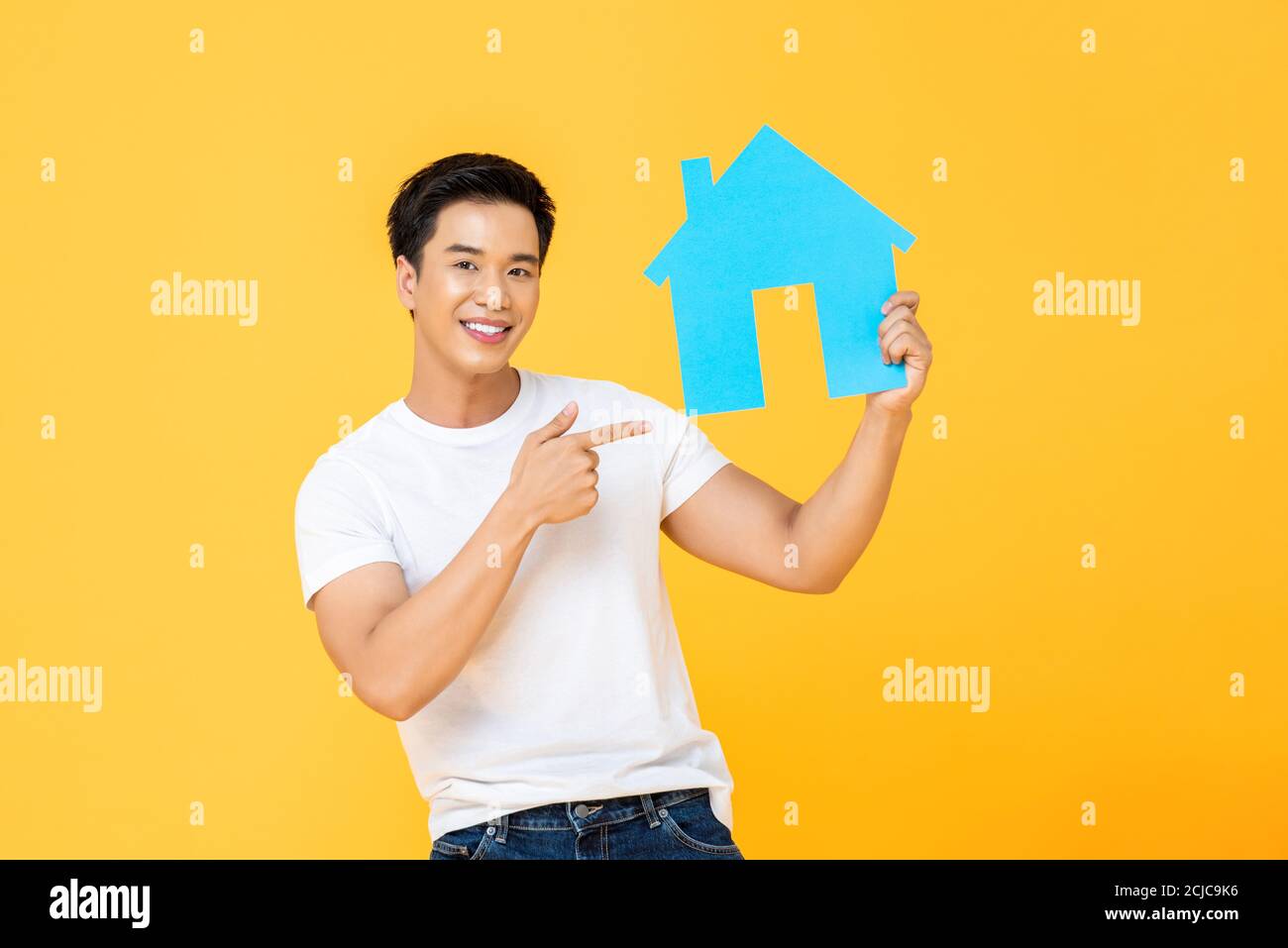 Schöner asiatischer Mann hält und zeigt auf Haus Ausschnitt isoliert Auf gelbem Studiohintergrund Stockfoto