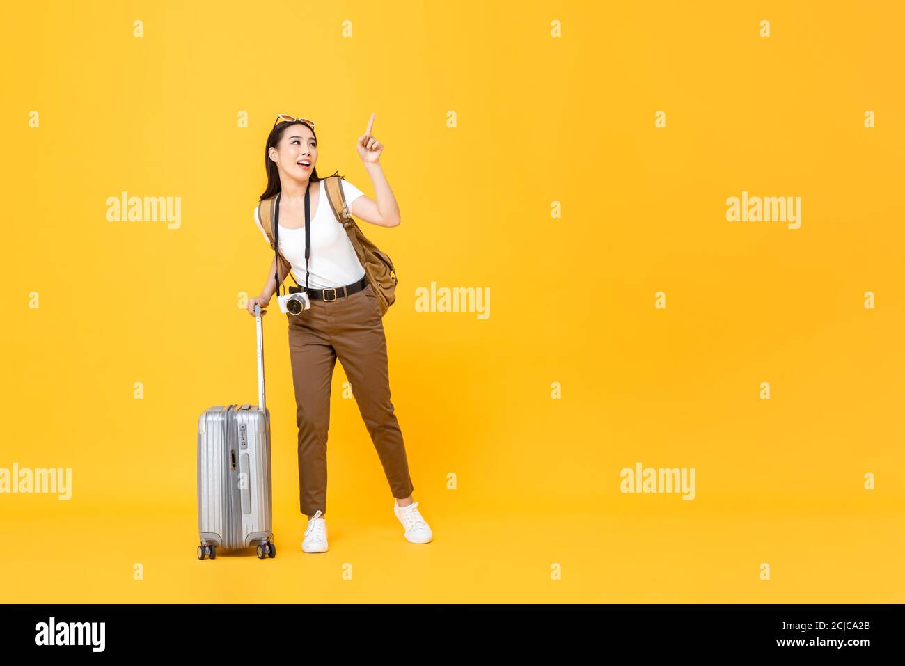 In voller Länge Porträt von niedlichen jungen asiatischen Frau Tourist poiting Hand up isoliert auf gelbem Studiohintergrund mit Kopierraum Stockfoto