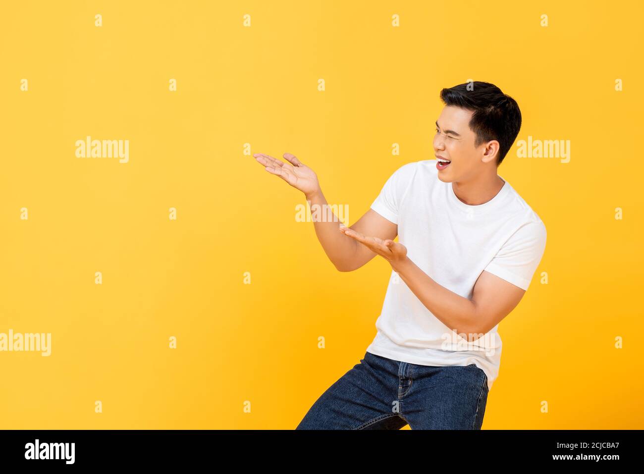 Fun Portrait von lächelnd junge gut aussehende asiatische Mann präsentiert mit Offene Handflächen Geste in isolierten Studio gelben Hintergrund mit Kopie Platz Stockfoto