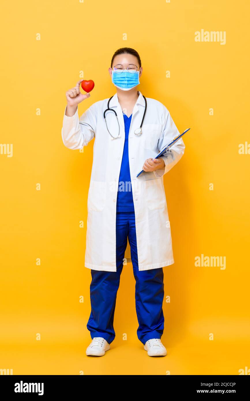 In voller Länge Porträt der jungen asiatischen Frau Arzt trägt Gesicht Maske mit einem roten Herzball in isoliertem Studio-Gelb Hintergrund Stockfoto