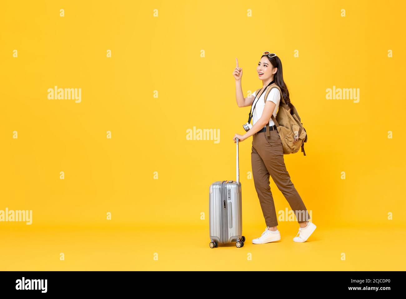 In voller Länge Reise Konzept Porträt von schönen lächelnden jungen asiatischen Frau Tourist mit Trolley-Tasche zeigt nach oben isoliert auf bunt Gelbes Studio-Bac Stockfoto