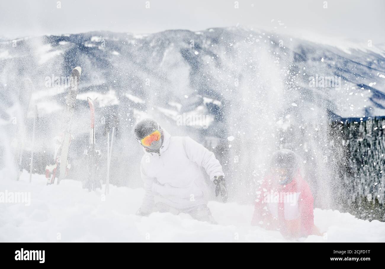 Zwei Skifahrer in Skianzügen und Helmen werfen frischen Pulverschnee hoch in die Luft. Mann und Frau in Skibrille Spaß im Skigebiet mit schönen Bergen im Hintergrund. Stockfoto