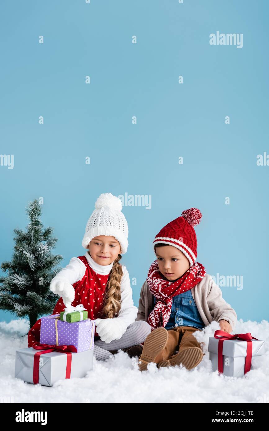 Kinder im Winteroutfit sitzen auf Schnee und schauen an Präsentiert neben Tanne isoliert auf blau Stockfoto