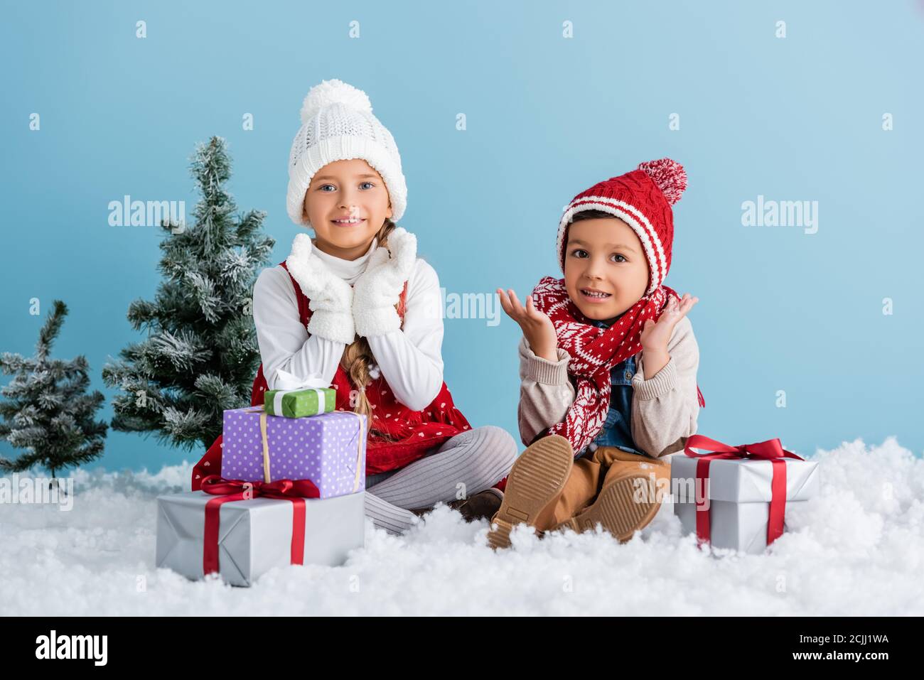 Kinder im Winteroutfit sitzen auf Schnee und gestikulieren in der Nähe Geschenke und Tanne isoliert auf blau Stockfoto