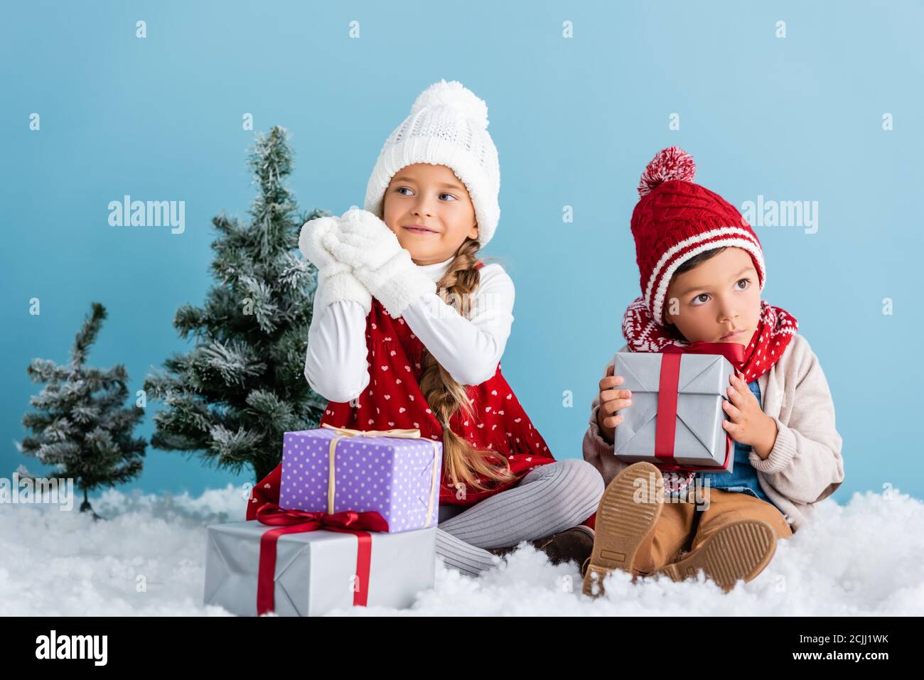 Kinder im Winter Outfit sitzen auf Schnee in der Nähe von Tannen und Präsentiert isoliert auf blau Stockfoto