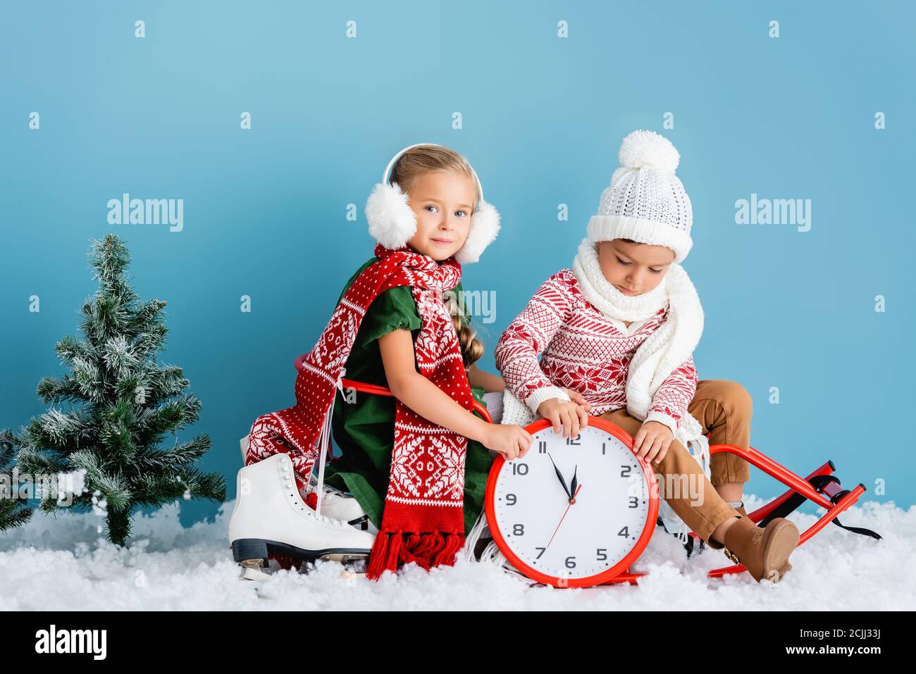 Kinder im Winter Outfit sitzen auf Schlitten in der Nähe Uhr, Kiefer und Schlittschuhe auf blau Stockfoto