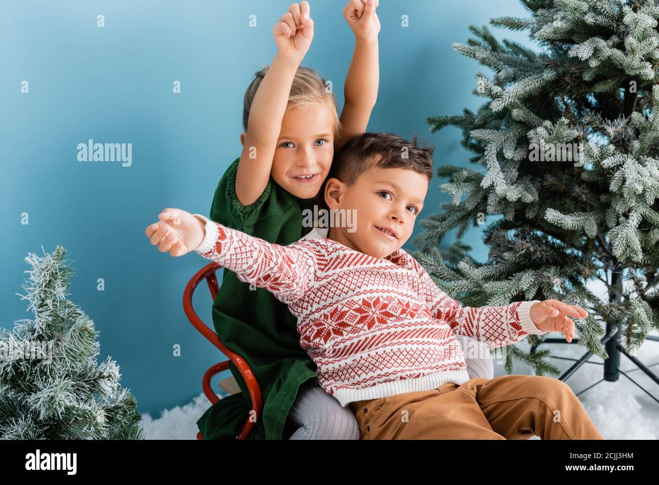 Aufgeregt Kinder mit Händen über Kopf sitzen in Schlitten in der Nähe Grüne Kiefern auf Blau Stockfoto