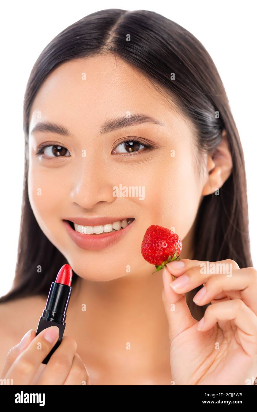 Junge asiatische Frau Blick auf Kamera, während halten roten Lippenstift Und frische Erdbeere in der Nähe Gesicht isoliert auf weiß Stockfoto