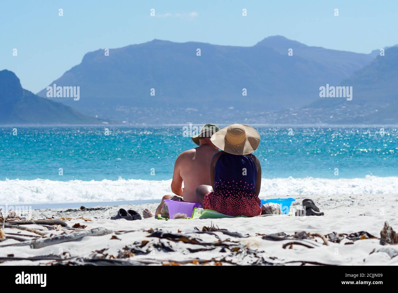 Ein Paar, kaukasischer Mann und schwarze Frau, sitzen zusammen auf weißem Sand und blicken auf den glitzernden blauen Ozean und die Berglandschaft Stockfoto
