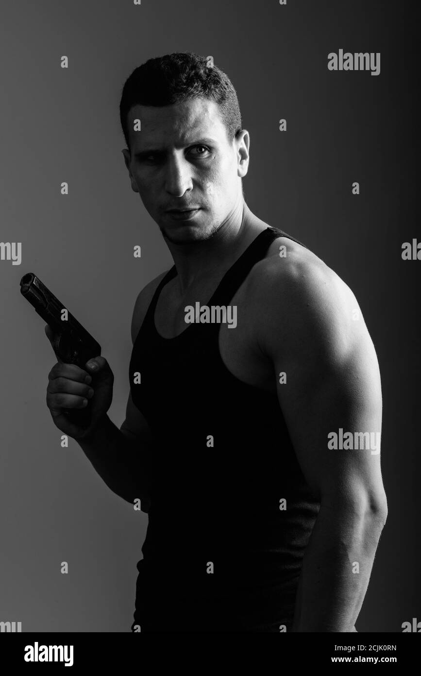 Nachdenklicher junger muskulöser persischer Mann, der Gewehr hält und zurückblickt Bereit für Action im dunklen Raum Stockfoto