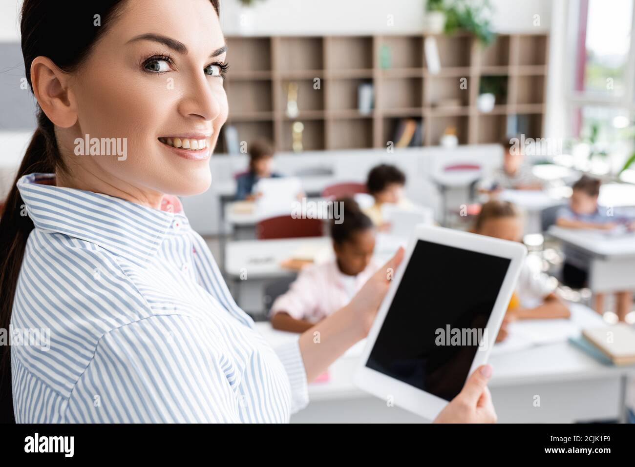 Selektiver Fokus des Lehrers mit digitalem Tablet mit leerem Bildschirm Und Blick auf die Kamera in der Nähe von multikulturellen Schülern im Klassenzimmer Stockfoto