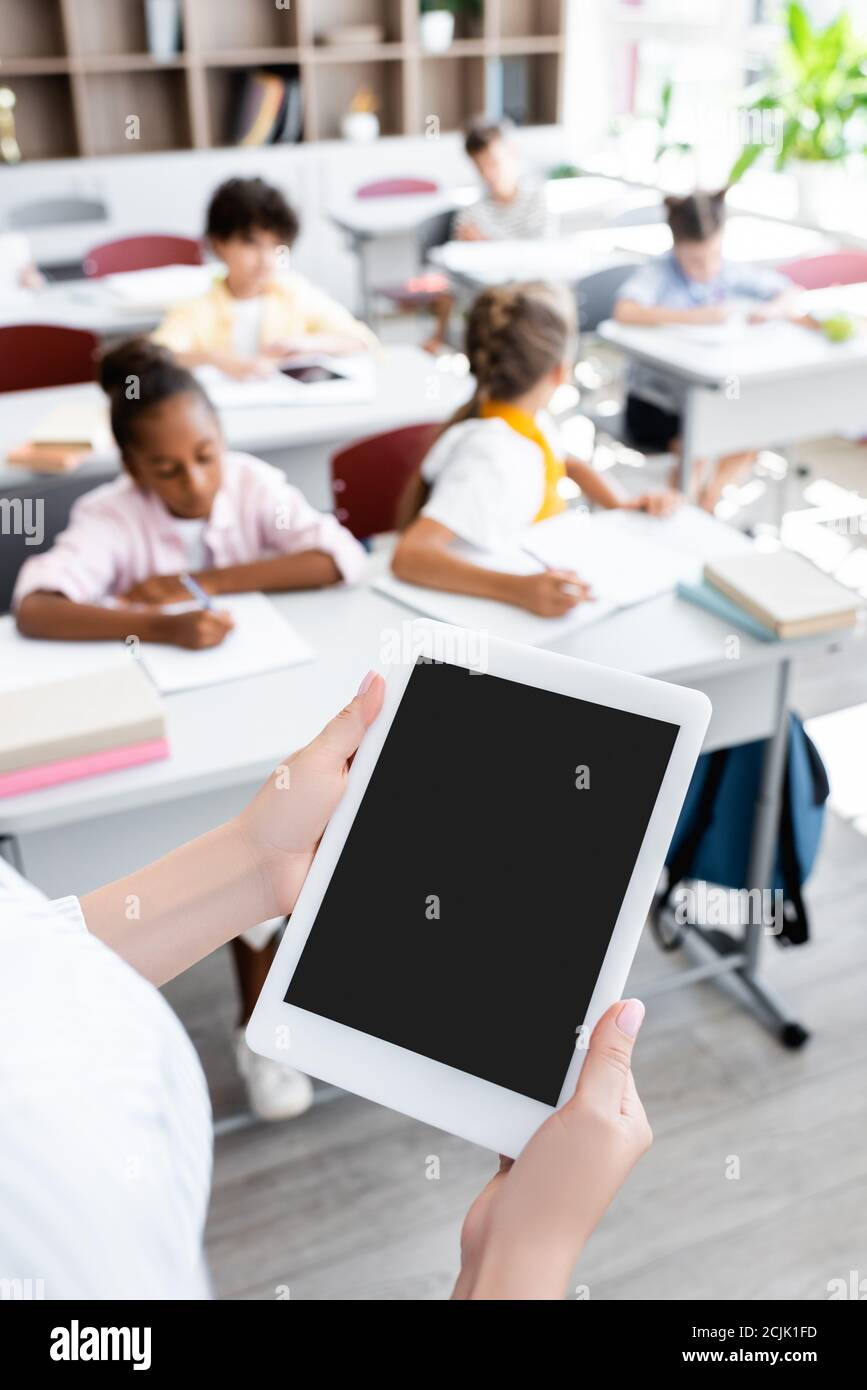 Teilansicht des Lehrers, der digitale Tablette mit leerem Bildschirm hält Nahe multikulturelle Schüler schreiben während des Unterrichts Stockfoto