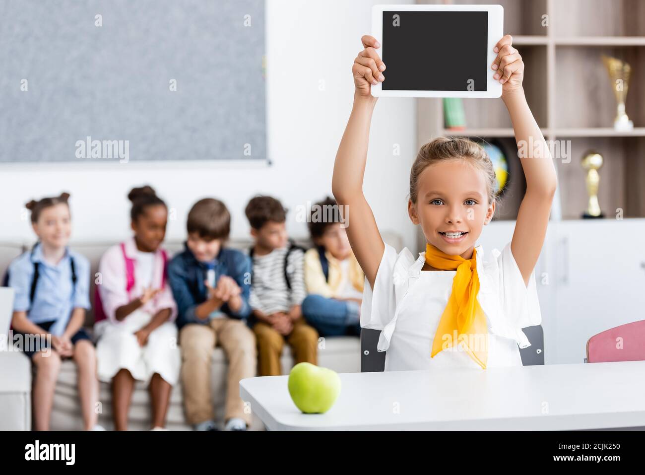 Selektiver Fokus von Schulmädchen zeigt digitale Tablette in der Nähe von Apfel auf Schreibtisch und multiethnische Klassenkameraden im Klassenzimmer Stockfoto