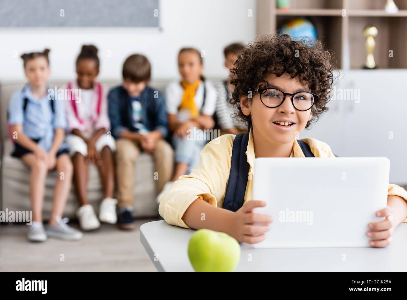 Selektiver Fokus des muslimischen Schuljungen mit digitalen Tablet in der Nähe von Apfel Auf dem Schreibtisch und multiethnische Klassenkameraden im Hintergrund Stockfoto