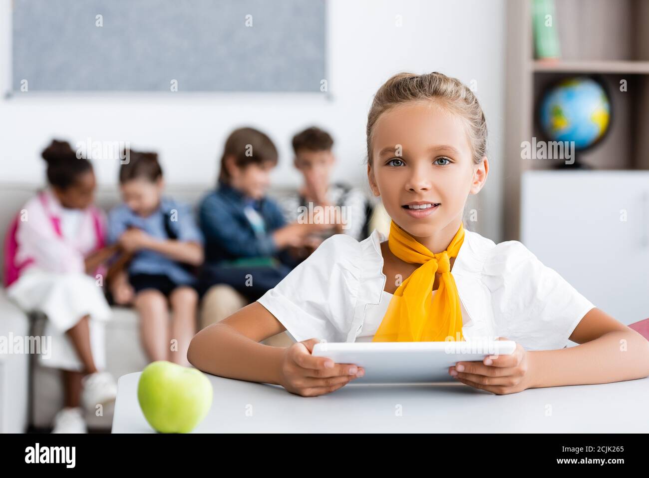 Selektiver Fokus der Schülerin hält digitale Tablette in der Nähe von Apfel auf Schreibtisch und multiethnische Freunde im Klassenzimmer Stockfoto