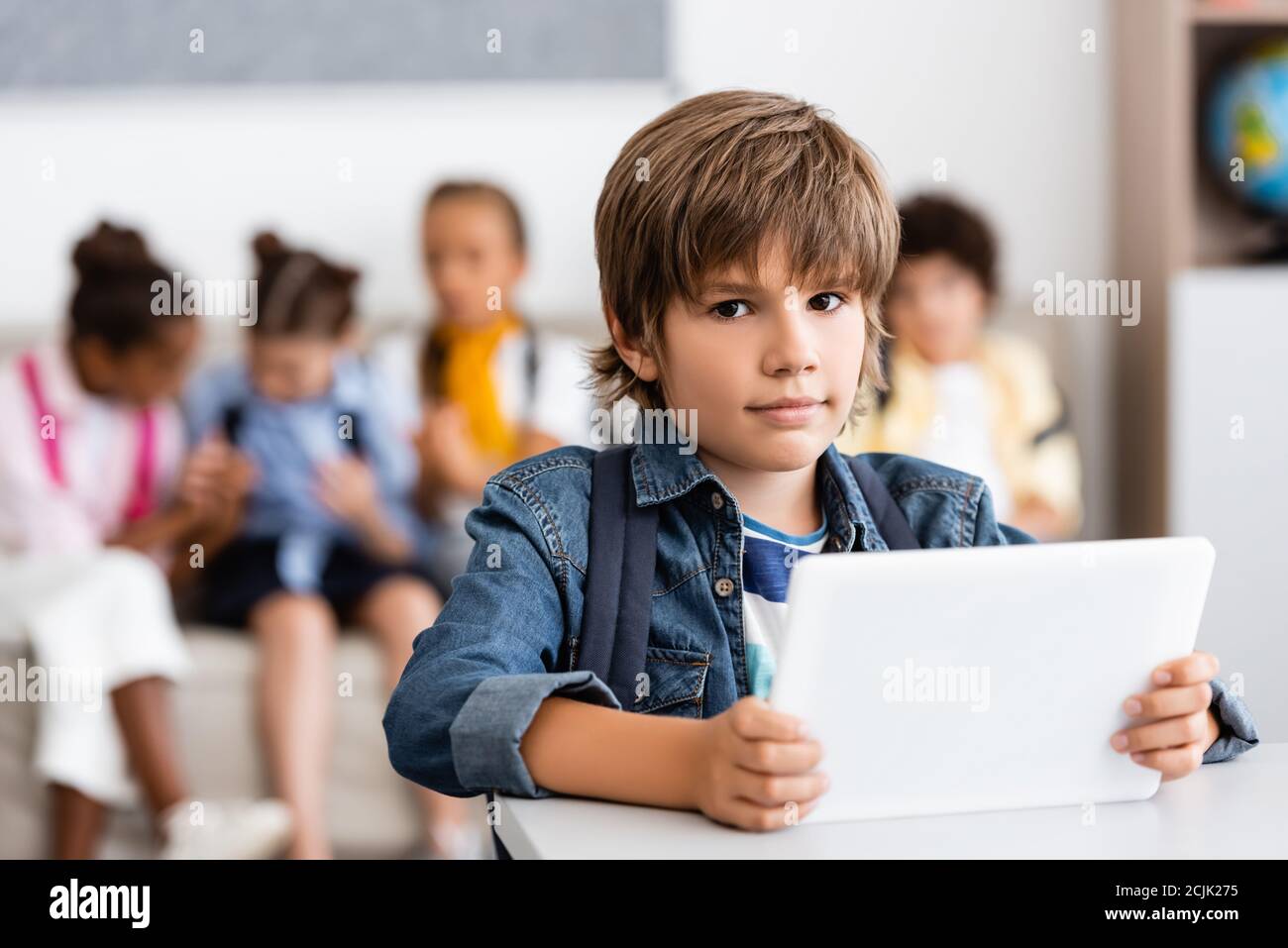 Selektiver Fokus des Schuljungen halten digitale Tablet und Blick auf Kamera am Schreibtisch im Klassenzimmer Stockfoto