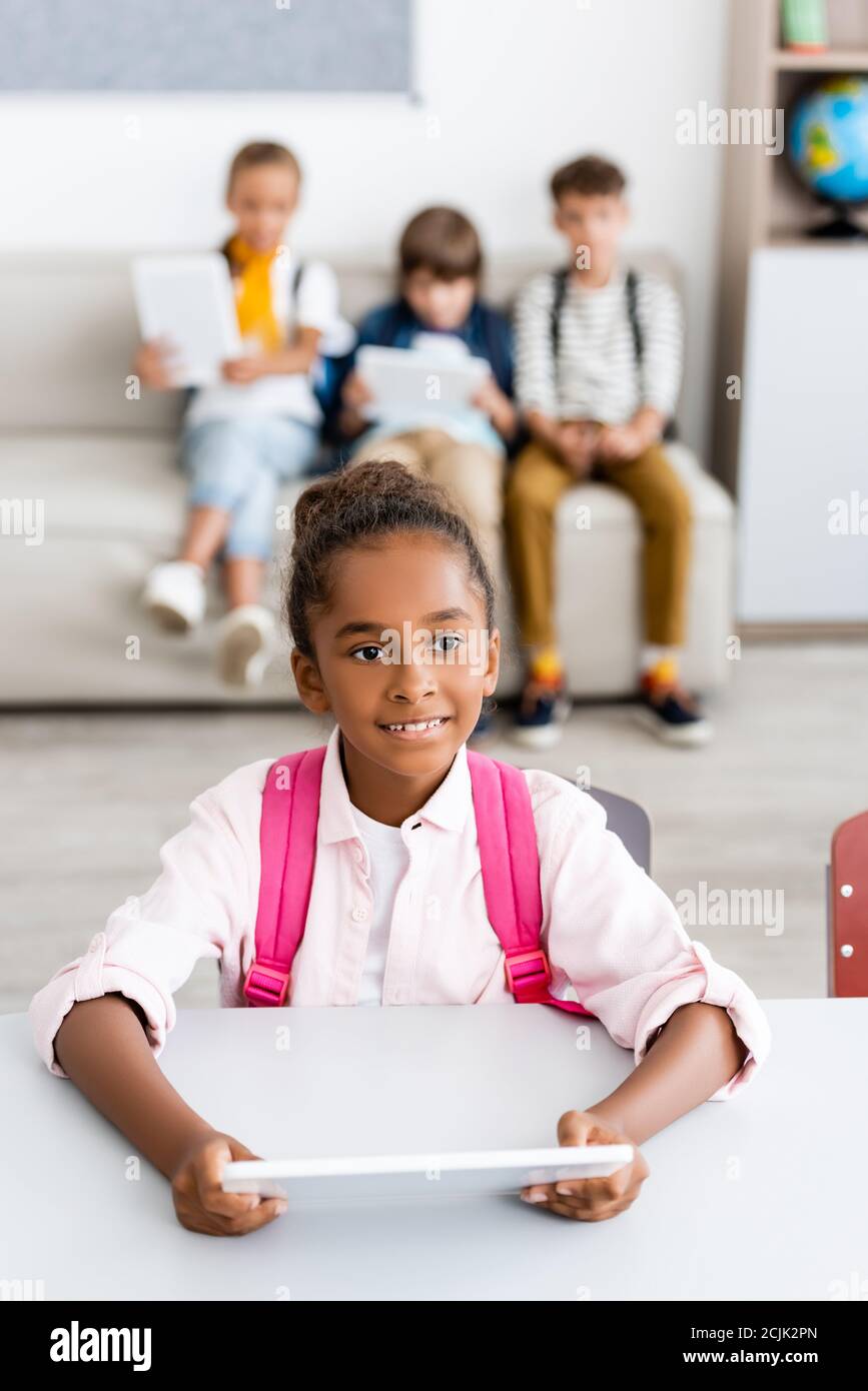 Selektive Fokus der afroamerikanischen Schulmädchen mit Rucksack halten digital Tablet am Schreibtisch in der Nähe von Freunden im Klassenzimmer Stockfoto