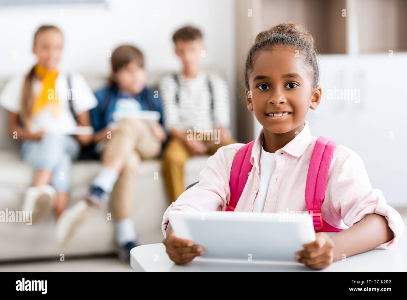 Selektive Fokus der afroamerikanischen Mädchen halten digitale Tablette an Schreibtisch mit Klassenkameraden im Hintergrund in der Schule Stockfoto