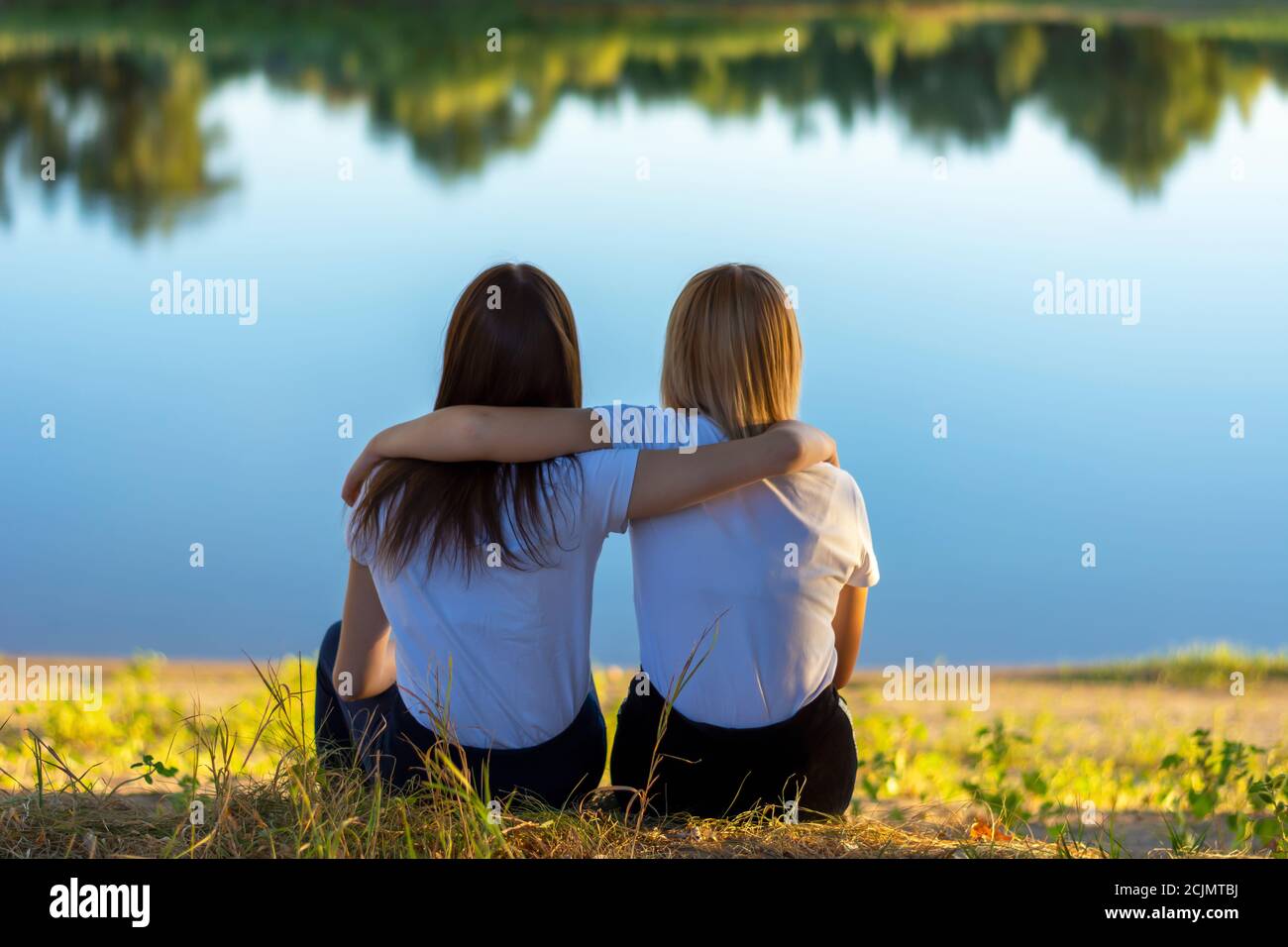 Zwei junge Frauen Rückansicht Umarmung beim Sitzen auf dem Gras in der Nähe des Flusses im Herbst. Freunde lehnten sich im Park aneinander. Stockfoto