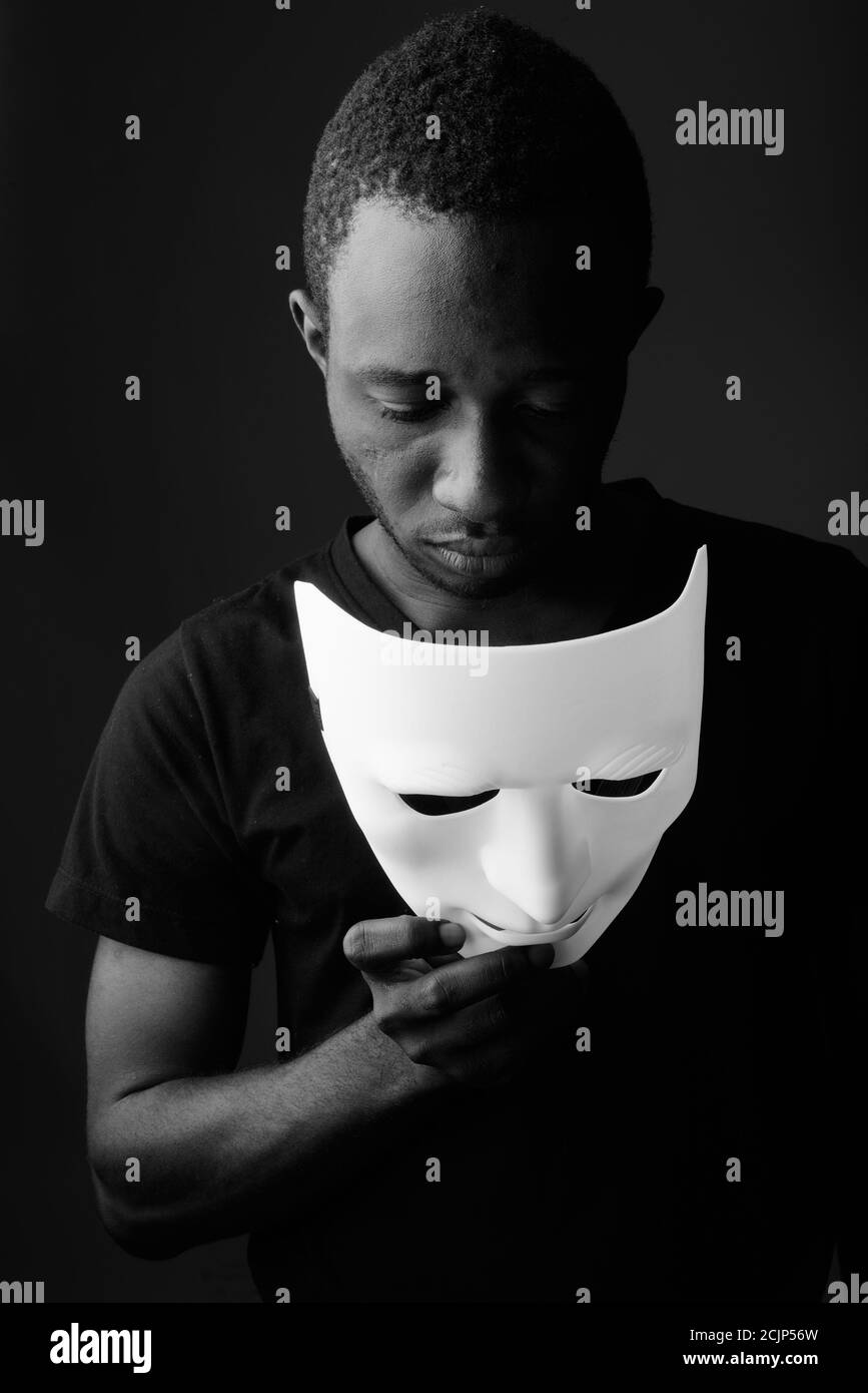 Studioaufnahme eines jungen schwarzen afrikanischen Mannes im dunklen Raum Maske halten und nach unten schauen Stockfoto