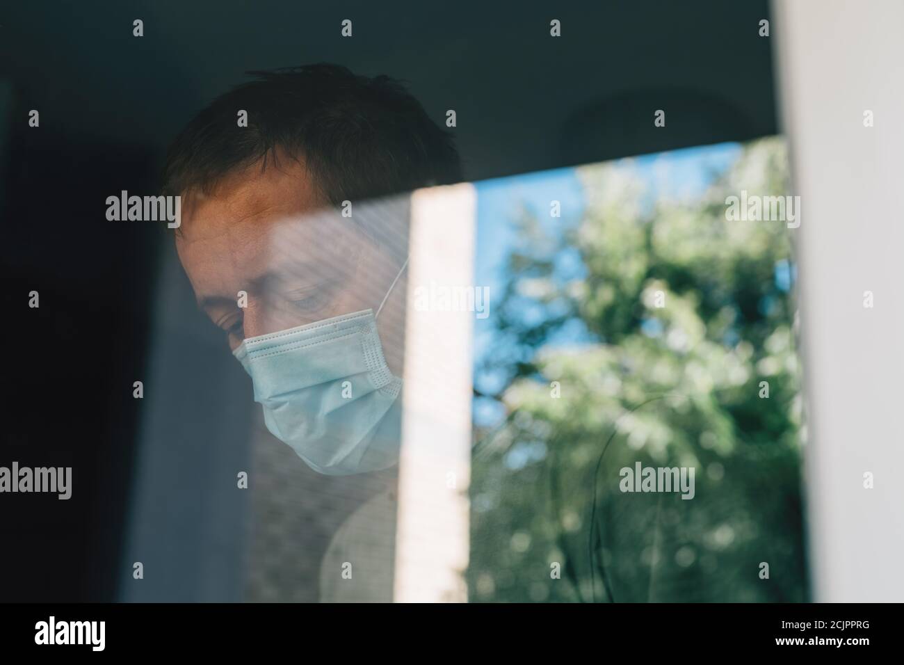 Besorgter Mann mit schützender Gesichtsmaske in Selbstisolation Hausquarantäne während Coronavirus Ausbruch, Porträt der erwachsenen männlichen Person hinter dem Fenster Stockfoto