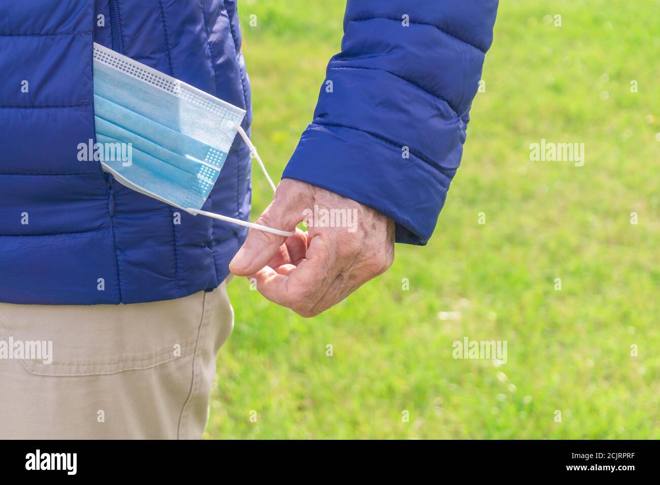 Alte menschliche Hand, die Schutzmaske aus der blauen Jackentasche herausnimmt. Senior Hand nimmt aus der Jacke Einweg medizinische Gesichtsmaske. Neues normales Leben. Stockfoto