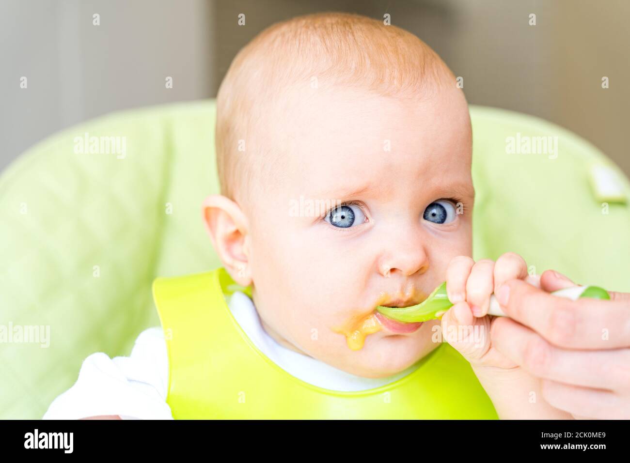 Ein kleines Kind mit blauen Augen und blonden Haaren sitzt in einem Hochstuhl und isst den ersten Köder. Gesunde Ernährung. Baby 6 Monate Stockfoto