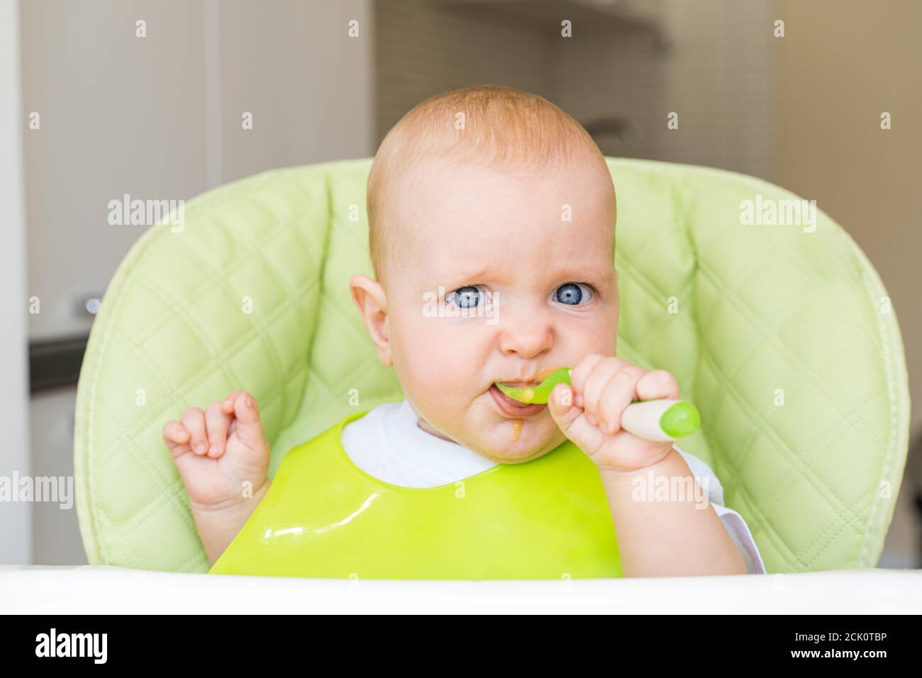 Ein kleines Kind mit blauen Augen und blonden Haaren sitzt in einem Hochstuhl und isst den ersten Köder. Gesunde Ernährung. Baby 6 Monate. Stockfoto