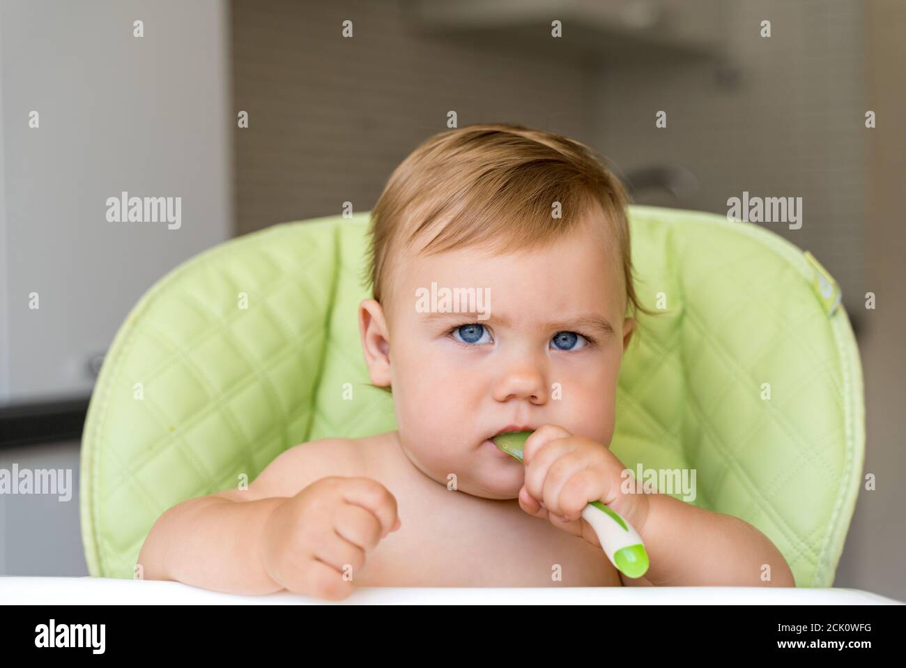 Ein kleines Kind mit blauen Augen und blonden Haaren sitzt in einem Hochstuhl und isst den ersten Köder. Gesunde Ernährung. Baby 11 Monate Stockfoto