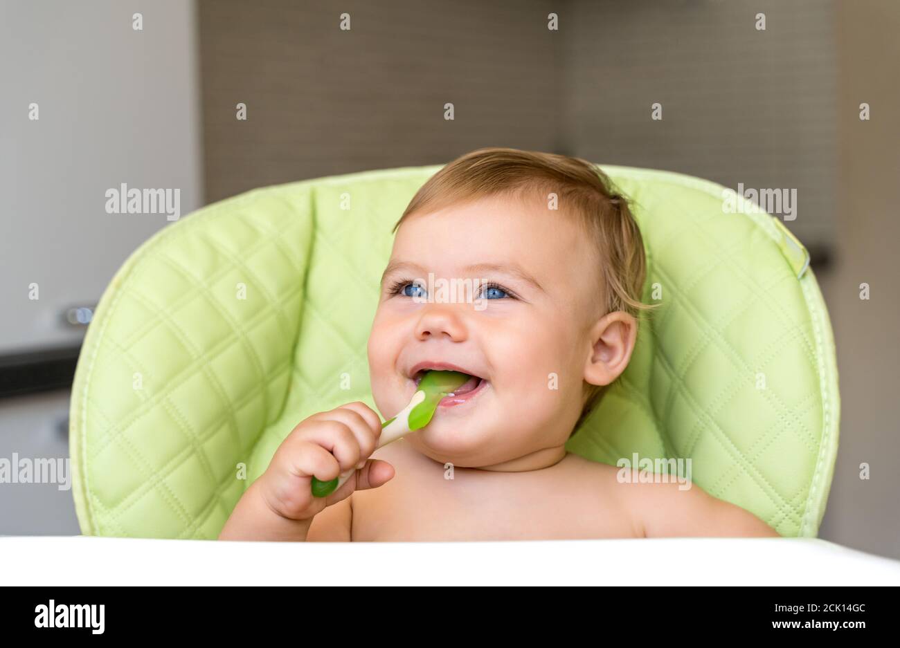 Ein kleines Kind mit blauen Augen und blonden Haaren sitzt in einem Hochstuhl und isst den ersten Köder. Gesunde Ernährung. Baby 11 Monate. Stockfoto