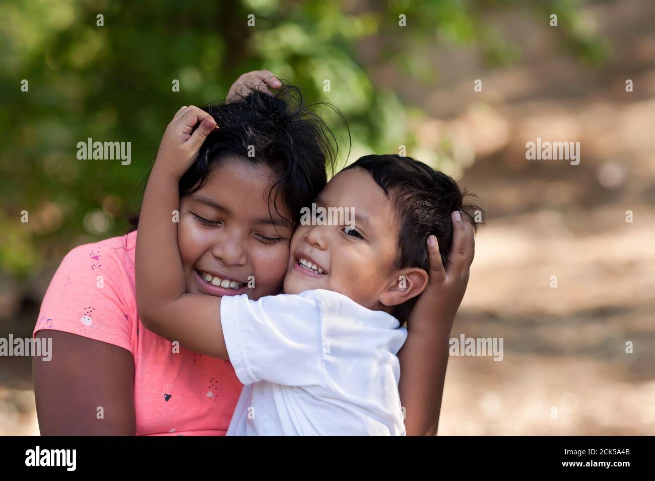 Der kleine Bruder umarmt seine ältere Schwester mit seinen Armen um ihr Gesicht und beide lächeln im Freien. Stockfoto