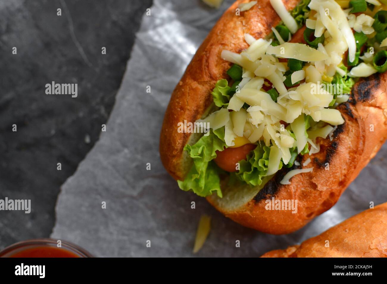 Ein Hot Dog mit Senf und Ketchup, Salat, Käse und Wurst. Dunkler Hintergrund. Lebensmittelhintergrund mit Kopierraum. Nahaufnahme. Draufsicht Stockfoto