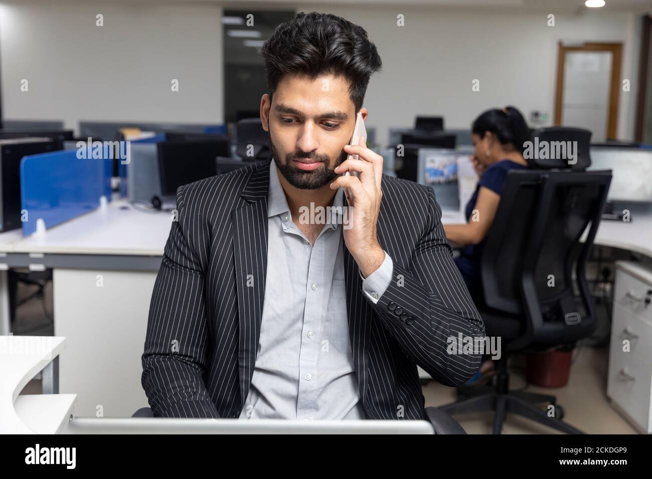 Porträt eines jungen indischen Geschäftsmannes, der am Telefon spricht, an seinem Arbeitsplatz vor Bürohintergrund sitzt, Unternehmensumgebung. Stockfoto