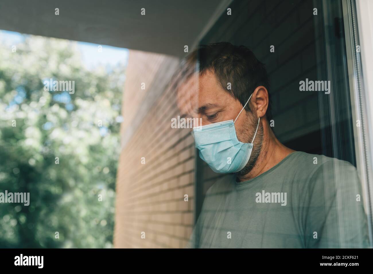 Besorgter Mann mit schützender Gesichtsmaske in Selbstisolation Hausquarantäne während Coronavirus Ausbruch, Porträt der erwachsenen männlichen Person hinter dem Fenster Stockfoto
