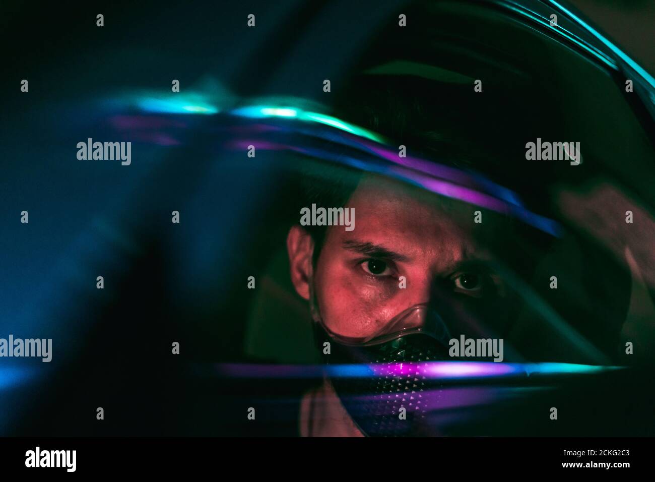 Ein junger Mann in einer Gesichtsmaske, der ein Auto fährt In der Nacht Stadt mit Neonlicht, die von der Glas seines Autos Stockfoto
