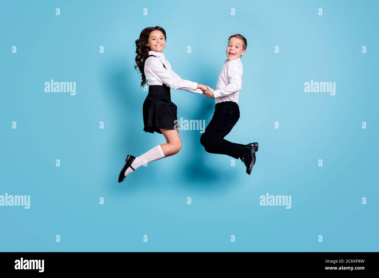 In voller Länge Profil Foto von zwei gegenüberliegenden kleinen Mädchen junge Schulkinder Bruder Schwester Klassenkameraden springen halten Hände feiern tragen weiß hemd schwarz Stockfoto