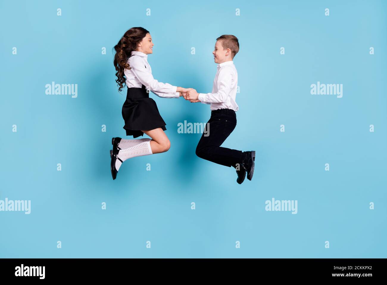 In voller Länge Profil Foto von zwei gegenüberliegenden kleinen Mädchen junge Schulkinder Bruder Schwester Klassenkameraden springen halten Hände mit Spaß tragen Weißes Hemd schwarz Stockfoto