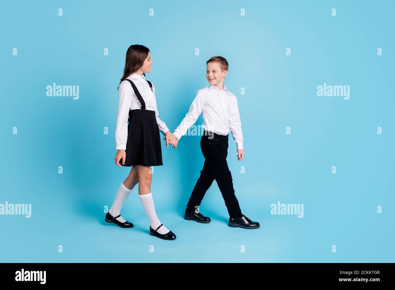 In voller Länge Profil Foto von zwei Mädchen junge Schulkinder lächeln Halten Sie die Hände Park zu Fuß nach der Schule tragen einheitliche weiße Hemd Schwarzes Kleid mit Hose Stockfoto