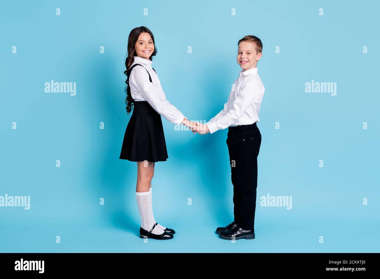 In voller Länge Profil Foto von zwei Mädchen junge Schulkinder glänzend Lächelnd halten Hände schauen Kamera warten Lehrer tragen uniform weiß hemd schwarz Hose Kleid Stockfoto