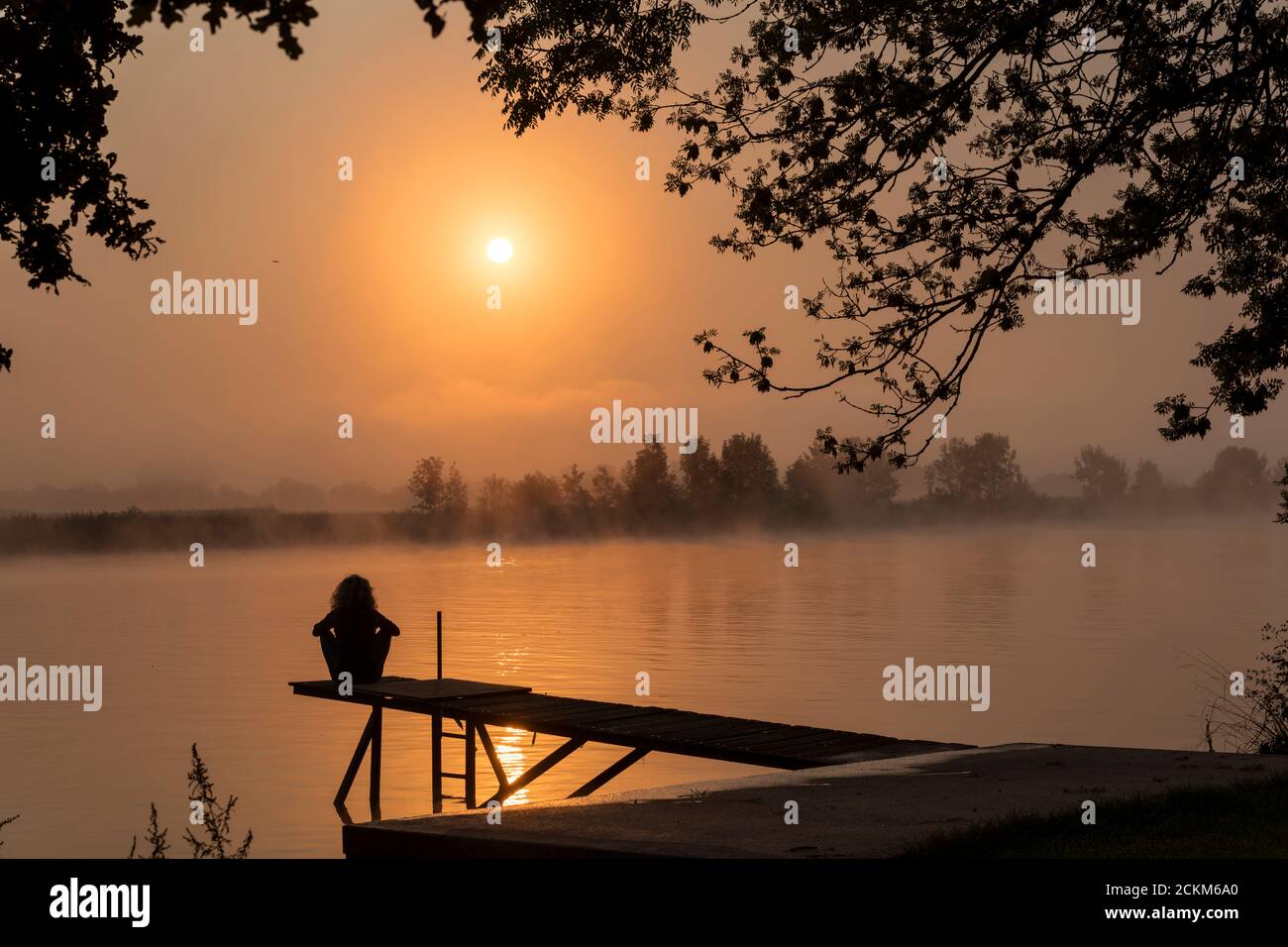 Silhouette Mädchen am hölzernen Steg während des Sonnenuntergangs in der frühen Morgen über dem Fluss maas in limburg in holland mit Die Bäume vernebeln und trüben Nebel Stockfoto
