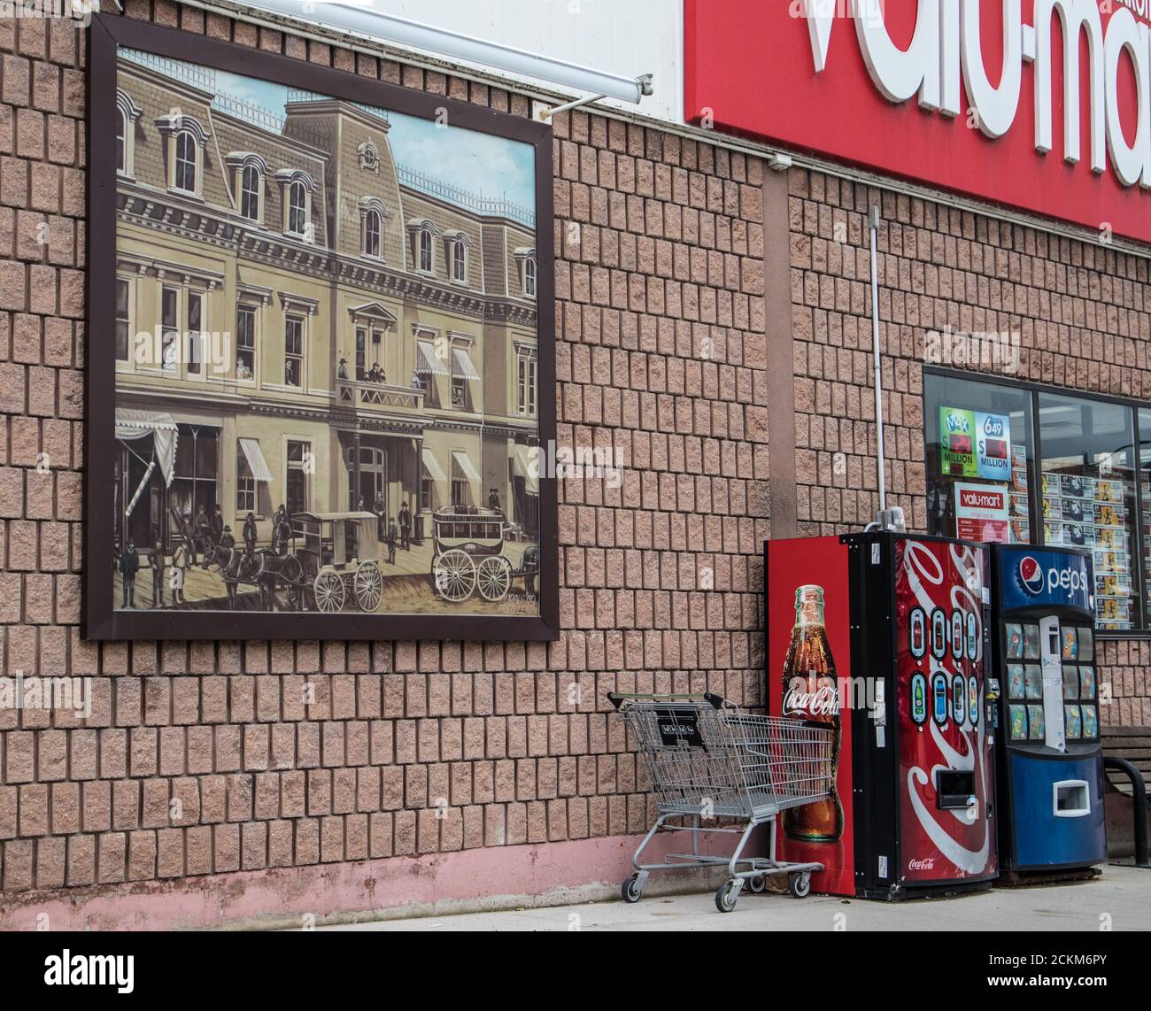 Ein Gemälde, das ein altes Gebäude darstellt, das ständig an der Wand des Valu-Mart, neben einigen Getränkeautomaten und einem Wagen ausgestellt ist. Stockfoto