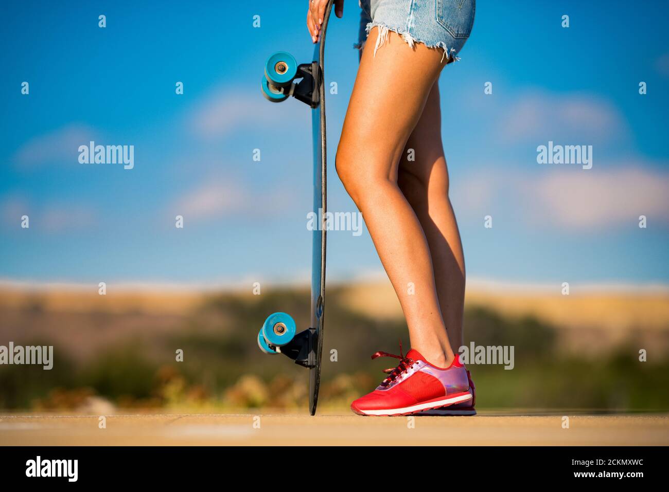 Stilvolle Passform und sportliche Frau mit schönen gebräunten Beinen und in roten Sneakers hält Longboard an glänzenden Sommertag. Aktivitäten im Freien. Straßenkultur. Stockfoto