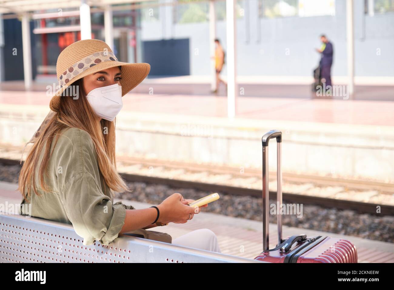 Die junge Frau trägt eine Gesichtsmaske und benutzt ihr Smartphone, während sie am Bahnhof auf den Zug wartet. Reisekonzept. Stockfoto