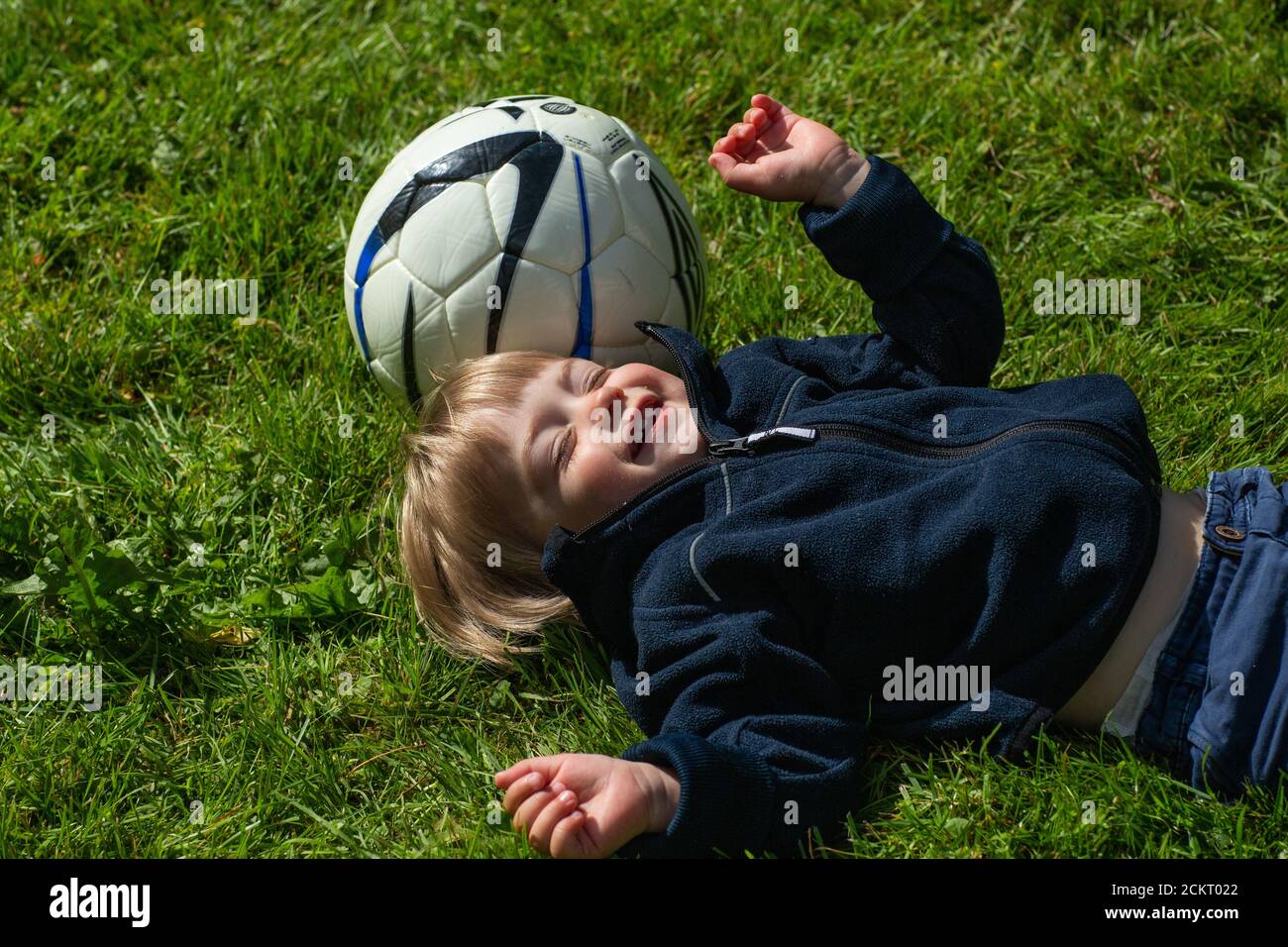 Glücklicher 2-jähriger Junge im Gras nebenan Ein Fußball Stockfoto