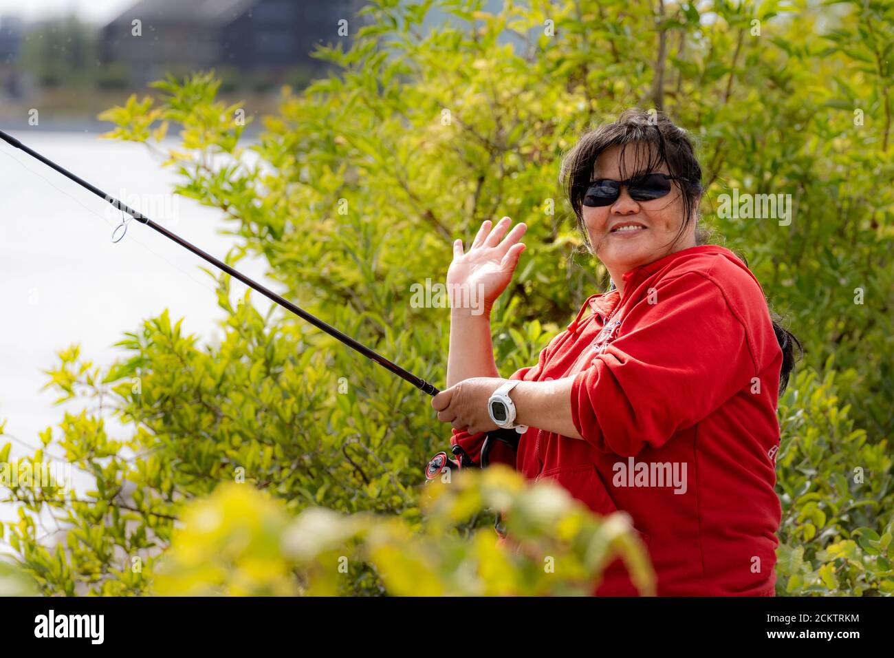 Eine lächelnde asiatische Frau mittleren Alters, in ihren 50ern, hält eine Angelrute und macht ein V-Zeichen. Grüne Büsche im Hintergrund Stockfoto