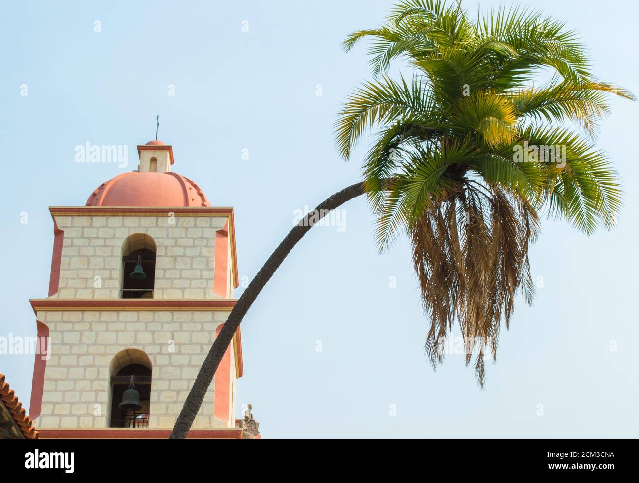 Eine anmutige, geschwungene Palme vor dem Glockenturm im spanischen Stil in der historischen Santa Barbara Mission in Kalifornien unter trübem Sonnenschein Stockfoto