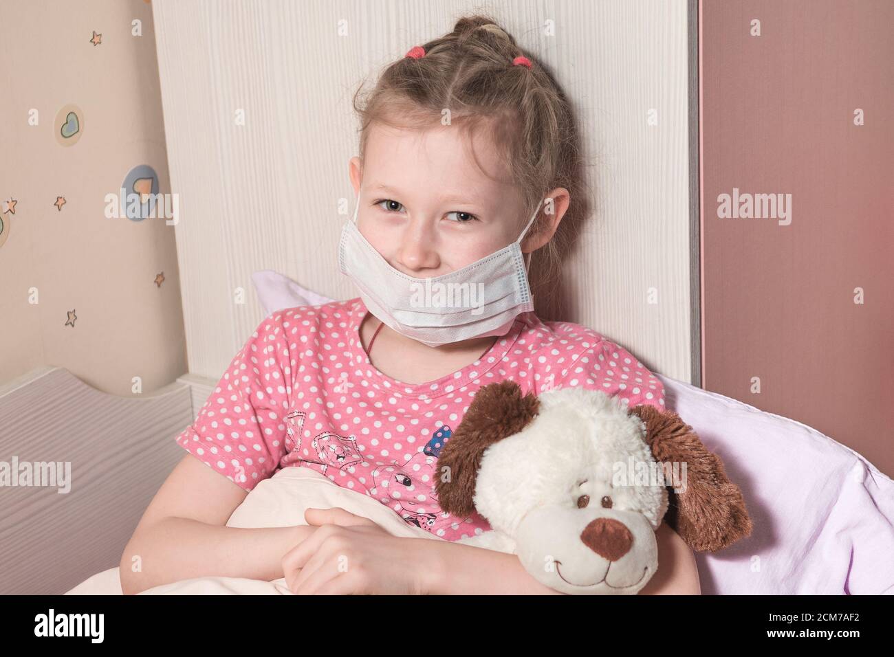 Baby Mädchen trägt Schutzmaske in Quarantäne, liegt im Bett mit einem weichen Spielzeug. Patient im Haus isoliert, um Infektionen zu verhindern. Coronovirus-Konzept. hor Stockfoto