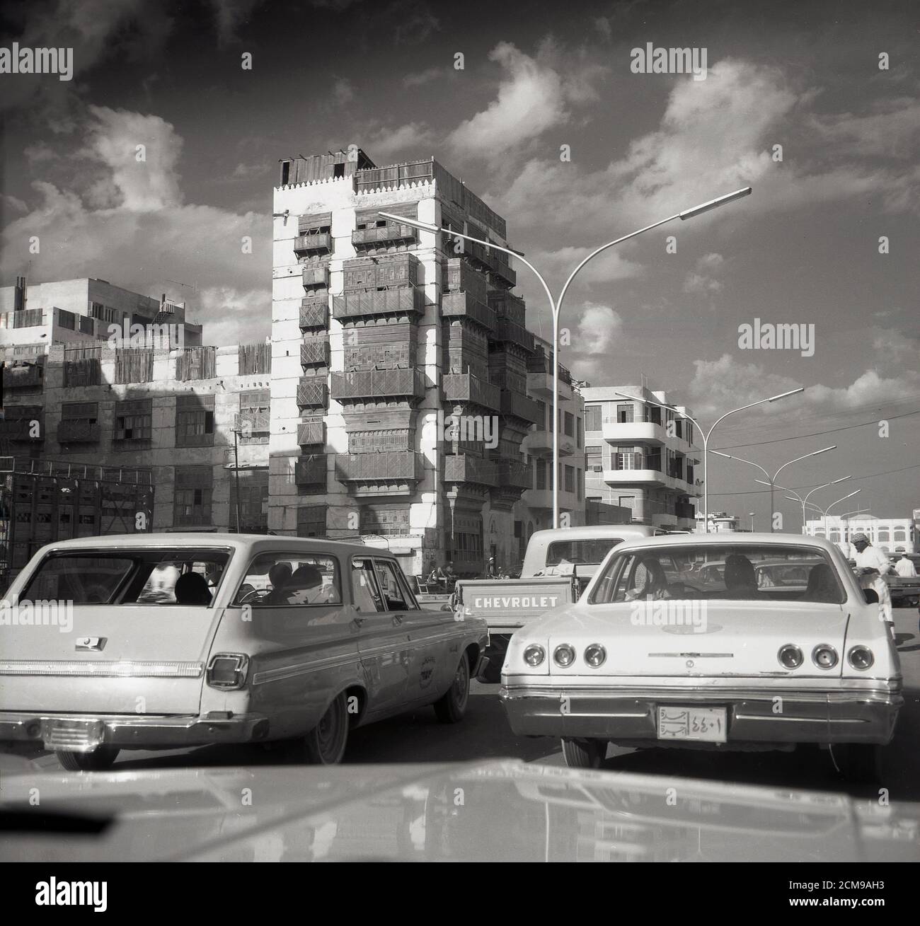 1960s, historisch von J Allan Cash zeigt breite amerikanische Autos der Zeit auf einer Straße in Jeddah, Saudi-Arabien, mit einem traditionellen saudischen Gebäude mit verzierten Holzbalkonen in der Ferne gesehen. Stockfoto